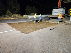 Night roadwork crew shoveling dirt near a bridge with an excavator in the background